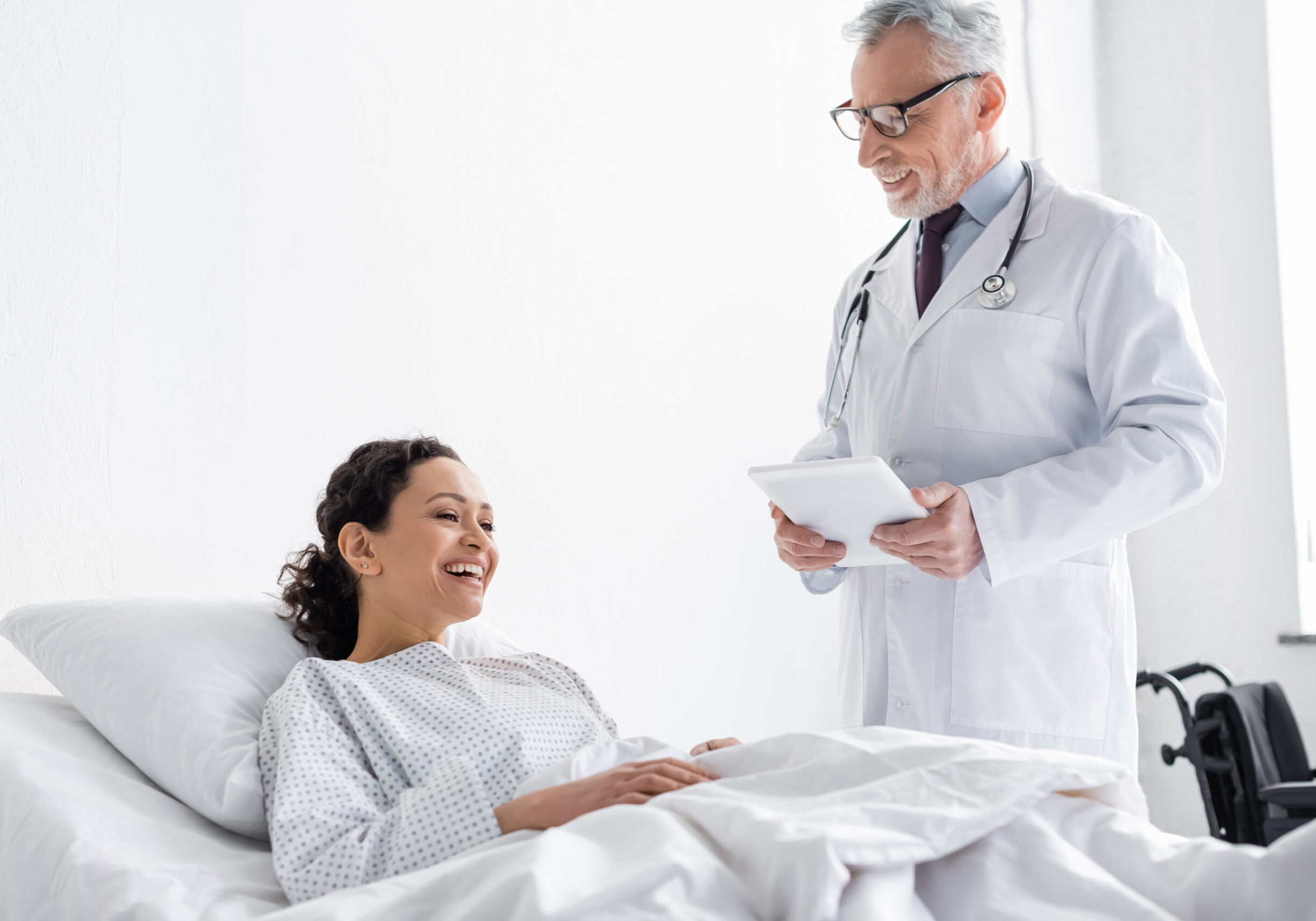 joyful african american woman lying in hospital bed near smiling doctor holding digital tablet